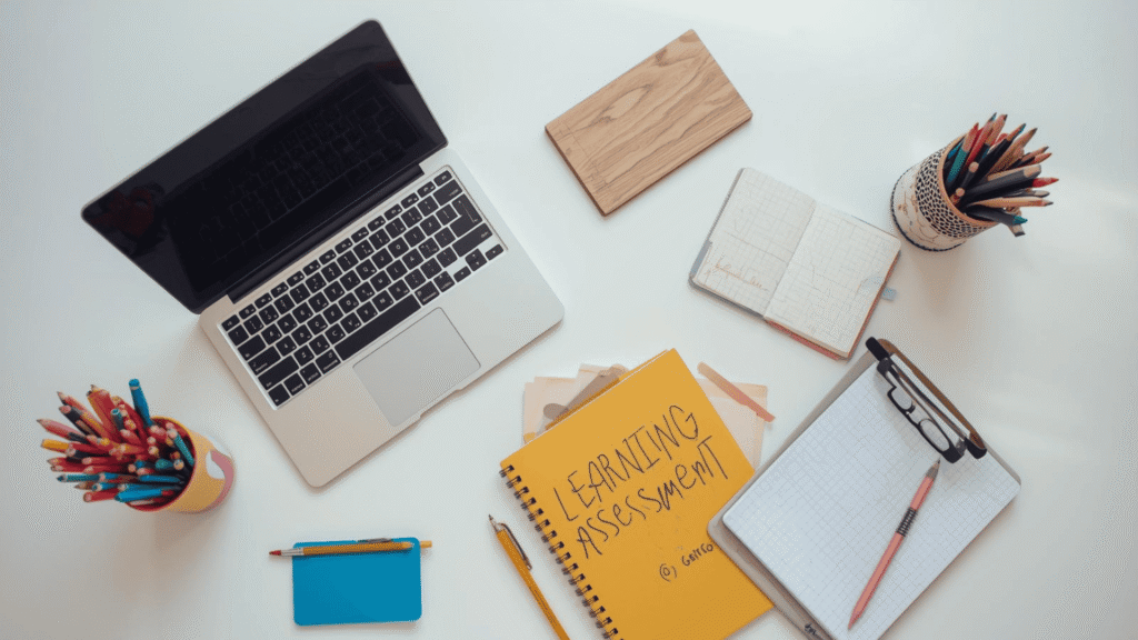 Meja kerja belajar dengan laptop, buku asesmen pembelajaran, alat tulis, dan buku catatan untuk perencanaan pembelajaran.