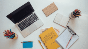 Meja kerja belajar dengan laptop, buku asesmen pembelajaran, alat tulis, dan buku catatan untuk perencanaan pembelajaran.